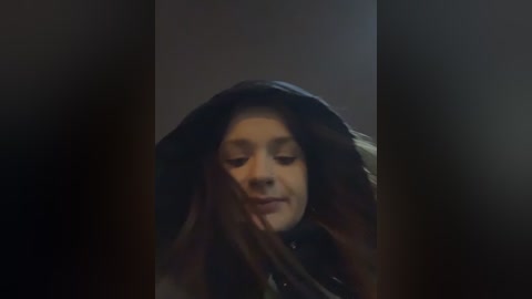 Video of a young woman with long, dark hair partially obscuring her face, wearing a black hoodie, in a dimly lit room with a dark background.