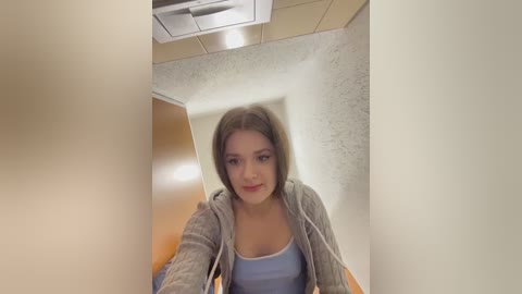 Video of a young woman with light skin, shoulder-length brown hair, and a light blue tank top, standing in a narrow, beige-walled hallway with a speckled marble floor.