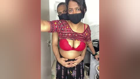 Video of a South Asian woman with medium brown skin and dark hair, wearing a black face mask, a red bra, and black pants, holding a man's hands on her stomach.