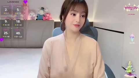 Video of a smiling East Asian woman with short brown hair in a beige robe, seated in a modern, minimalistic room with a plush teddy bear and a calendar showing 18% humidity.