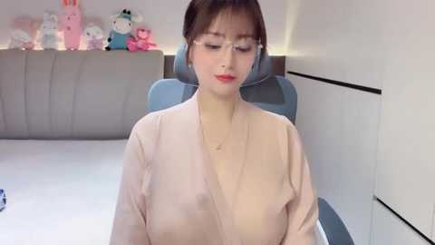 Video of an Asian woman with short brown hair and glasses, wearing a light pink robe, seated on a blue gaming chair in a modern room with white walls and plush toys.