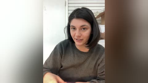 Video of a woman with straight, shoulder-length black hair, wearing a gray sweater, sitting indoors. A person's hand is visible in the right foreground.