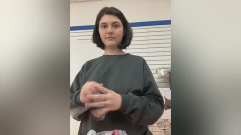 Video of a young woman with short black hair, wearing a grey sweatshirt, standing in an office with white blinds and a blue stripe.