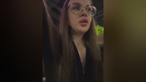 A dimly-lit video of a young woman with fair skin, long brown hair, and round glasses, wearing a black jacket and a necklace, sitting indoors with blurred background.