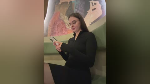 Video of a young woman with long dark hair, glasses, and a black dress, holding a smartphone, standing in front of a colorful mural.