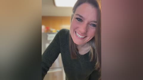 Video of a young Caucasian woman with light skin, straight brown hair, and blue eyes, smiling while leaning forward from a hallway into a kitchen. She wears a dark green sweater and stands in a modern kitchen with stainless steel appliances and wooden flooring.