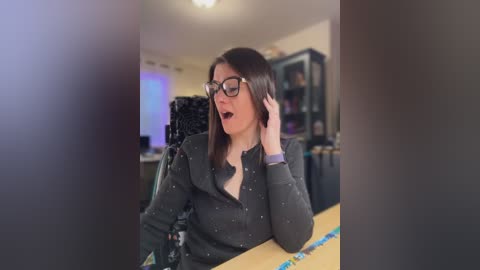 Video of a woman with shoulder-length brown hair, wearing black-framed glasses, a gray sweater, and a blue bracelet, holding a phone, in a cluttered office with shelves and a computer monitor.
