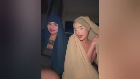 Media: Video of two women in a dimly lit room. The woman on the left has a dark blue hijab and red lipstick, smiling. The woman on the right has a beige hijab and red lipstick, holding her head in a playful manner.