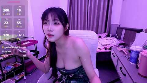 Video of an Asian woman with long black hair, wearing a strapless camo top, sitting at a messy desk in a dimly-lit room with purple lighting.