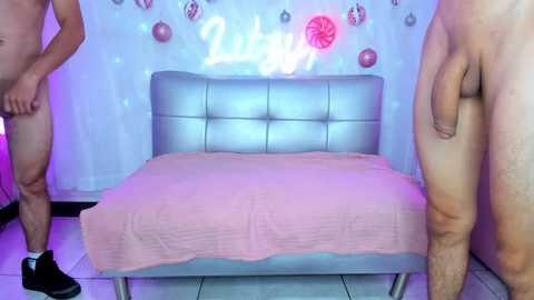 Video of two naked men, one holding a flaccid penis, standing near a pink-covered bed in a light blue room with \"2019\" decor.