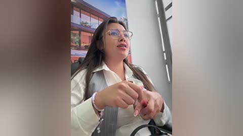 Video of a middle-aged woman with long dark hair, wearing glasses, a white blouse, and a black purse, gesturing with both hands, in an office with a modern mural background.