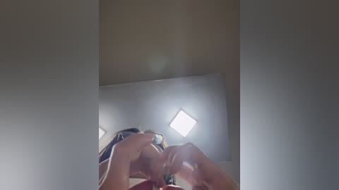 Video of a woman with long, straight hair, wearing a pink top, covering her face with her hands. The image is dimly lit with bright light coming from the top left corner.