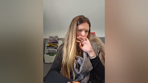 Video of a young woman with long blonde hair, wearing a grey scarf and black sweater, holding her nose, standing in a cluttered room with magazines and a fur coat on a chair.