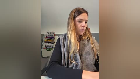 Video of a young woman with long, straight, blonde hair, wearing a black long-sleeve top and a gray, fluffy cardigan. She sits at a desk in a brightly lit room with a \"St. Louis Post-Dispatch\" newspaper rack and other magazines in the background.