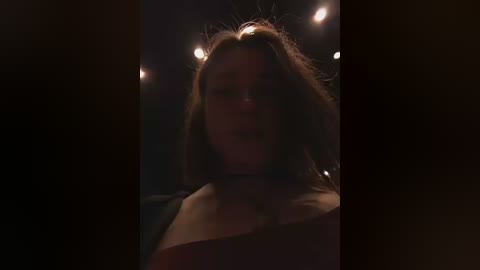 Video of a young woman with shoulder-length, tousled brown hair, wearing a black choker, and a dark top, illuminated by dim, soft lighting, creating a moody, intimate atmosphere.