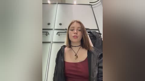 Video of a young Caucasian woman with light skin, shoulder-length blonde hair, wearing a black jacket over a red tank top, standing in a modern, brightly lit dressing room with mirrored walls and metal racks.