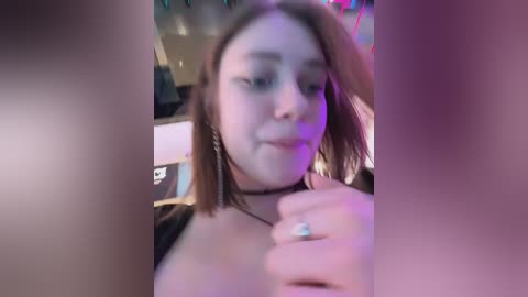 A blurry video of a young woman with straight, shoulder-length brown hair, wearing a black top and silver hoop earrings, taken in a dimly lit room with neon lighting.