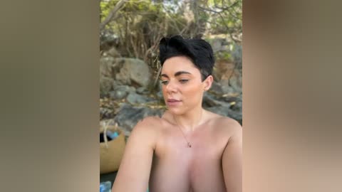 Video of a fair-skinned, topless woman with short dark hair, standing in a natural, rocky, outdoor setting with sparse greenery.