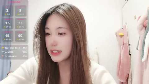 Video of a young Asian woman with long, straight brown hair, fair skin, and a light pink lip color, wearing a white top, in a modern, well-lit room. A digital screen displays health metrics in the background.