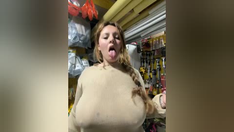 Video of a woman with large breasts, wearing a tight beige sweater, sticking out her tongue in a playful manner. Background shows a cluttered hardware store with shelves and hanging items.
