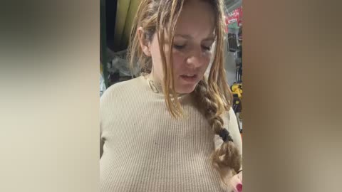 Video of a fair-skinned woman with long, blonde hair in a braid, wearing a cream-colored ribbed sweater, standing indoors in a dimly lit room with cluttered background.