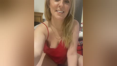 Video of a smiling, light-skinned woman with long blonde hair, wearing a red lace lingerie top, sitting on a bed in a bedroom.