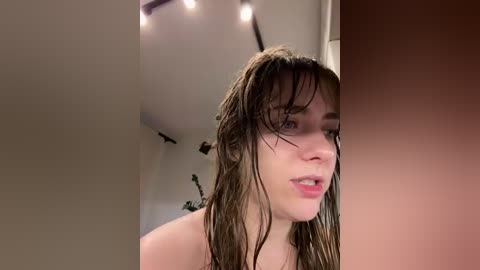 Video of a young woman with wet, messy hair, pale skin, and light makeup, looking slightly surprised. She stands in a bathroom with a beige wall and a blurred background, under bright lights.