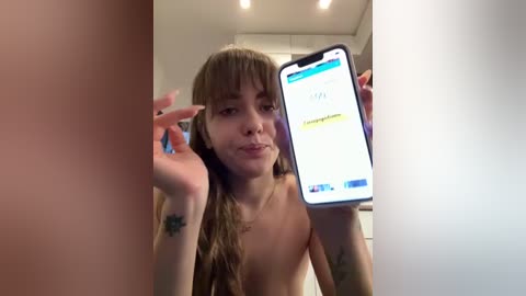 A topless, fair-skinned woman with long, brown hair takes a selfie, holding a phone showing a text message. She has tattoos on her arms. The background is a white room with recessed lighting.