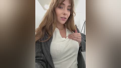 Video of a young woman with long, wavy brown hair, fair skin, and light makeup. She wears a cream ribbed top under a black blazer, holding a phone and smiling. Background shows a modern room with white walls and soft lighting.