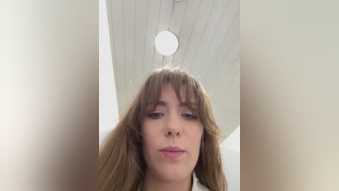 A video of a young woman with long, light brown hair, bangs, and fair skin, standing indoors with a white ceiling and recessed lighting.