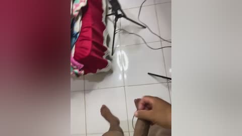 A video shows a woman's legs on a tiled floor, wearing a red dress, with a pair of black shoes and a white purse nearby, against a pink wall.