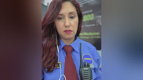 Video of a Latina police officer with long, wavy red hair, wearing a blue uniform with a red tie, holding a radio, standing in a blurred, dimly lit room with a clock displaying 8:00.