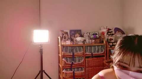 Video of a young girl with light brown hair, wearing a pink shirt, looking towards a bright light on a tripod. Background features a wicker bookshelf with various books and a stuffed animal.