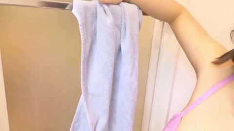 A video of a person with light skin and straight brown hair, wearing a pink bra, holding a blue towel in a beige-tiled bathroom.