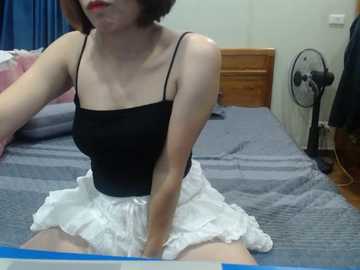 Video of an Asian woman with short brown hair, wearing a black spaghetti strap top and white ruffled skirt, sitting on a bed with blue sheets in a dimly lit bedroom.