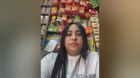 Video of a Latina woman with long black hair and medium skin tone, wearing a white \"CASA MIA\" t-shirt, standing in a brightly lit grocery store aisle filled with colorful packaged snacks.