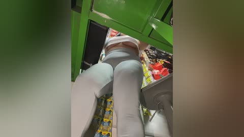 A video of a woman's legs in light blue tights and high-heeled shoes, standing in a bright green, well-lit room with shelves filled with colorful products and boxes.