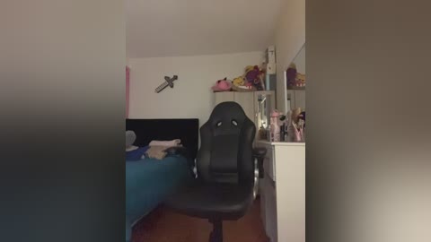 Video of a minimalist bedroom with a black gaming chair facing a bed with blue sheets. A white dresser holds toys and a mirror, while a wall-mounted cross adds a religious touch.