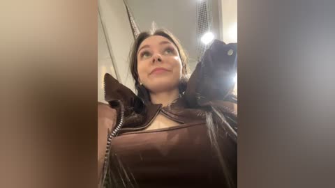Video of a woman with short, dark hair, wearing a brown leather jacket, taking a selfie in a dimly lit room with a reflection on a mirror.