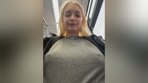 Video of a young, fair-skinned woman with blonde hair, wearing a grey T-shirt and black jacket, sitting in a train. Background features grey seats and metal poles.