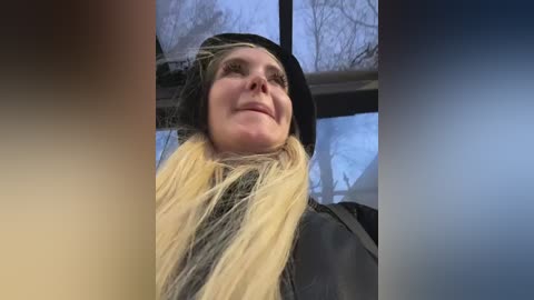 A video of a smiling woman with long blonde hair, wearing a black hat, standing in front of a large window, with a blurred background of bare trees outside.