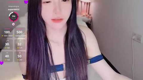 Video of a young, fair-skinned woman with long, straight black hair, wearing a black harness and a white tank top. She is indoors, with a bed and lamp in the background.