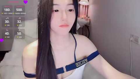 Video of an Asian woman with long black hair, fair skin, and a petite frame, wearing a white bra and blue strapless harness, in a bedroom with a white bed, pink lamp, and heart icons on the screen.