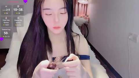 A video of a young Asian woman with long black hair, pale skin, and a slender build, gently plucking her eyebrows while sitting on a white chair in a modern, softly lit room.
