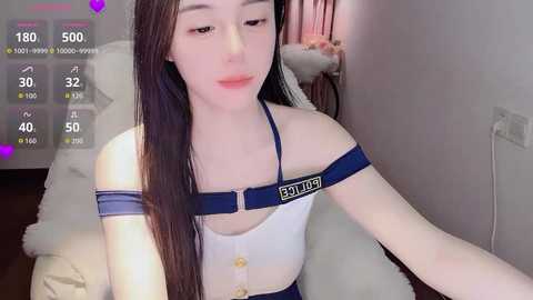 A video of a young Asian woman with long black hair, wearing a white dress with blue straps, sitting on a bed in a dimly lit room.