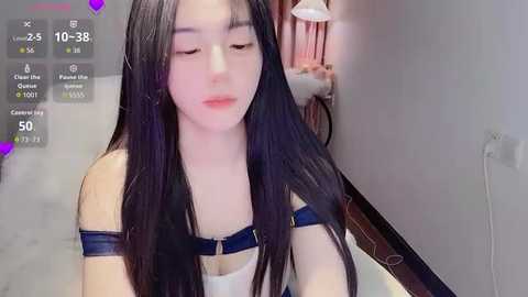 Video of an Asian woman with long, straight black hair and pale skin, wearing a blue off-shoulder top, sitting in a dimly lit room with a lamp and pink curtains.