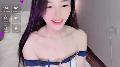 Video of a smiling, fair-skinned, petite Asian woman with long black hair, wearing a blue off-shoulder top, in a simple indoor setting.