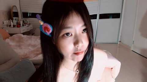 Video of an East Asian woman with long black hair and a red hat adorned with a colorful flower, smiling. She wears a white top and sits on a plush bed in a bright, modern bedroom.