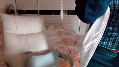 Video of a white leather sofa with a fluffy pink blanket, partially covered by a teal jacket, set against white blinds in a cozy, well-lit room.