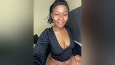 Video of a smiling, medium-dark-skinned woman with shoulder-length black hair, wearing a black, long-sleeved crop top and beige pants, sitting on a dark gray couch.